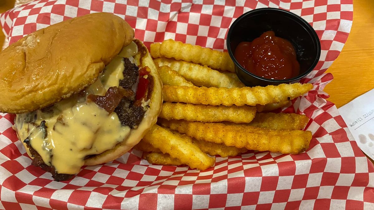 A smash burger with melted cheese and bacon, crinkle fries and a ketchup cup in red-checkered paper