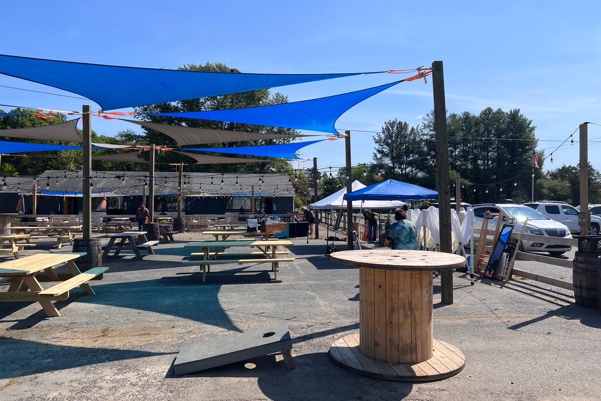 The patio under blue and grey shade sails: picnic tables, a cable-spool table, a cornhole board, the pavilion behind