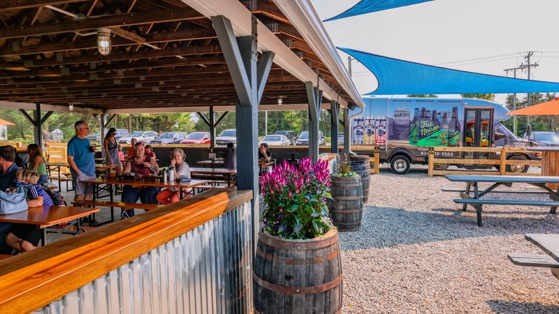 The pavilion bar with a corrugated-metal front and a barrel of magenta flowers, guests at tables, a wrapped shuttle bus parked behind